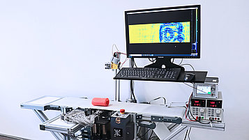 Das Foto zeigt den FBH Terahertz-Linienscanner im Betrieb. Auf dem Laufband liegt eine Kunststoffrolle, in die ein Tesafilmroller eingewickelt ist. Dieser lässt sich auf dem darüber liegenden Monitor erkennen.