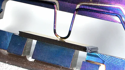 Close-up of a thin stacked wafer held between metal clamps with iridescent blue and purple reflections on the surrounding metallic surfaces.