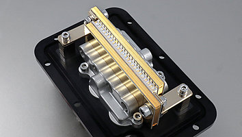 Das Bild zeigt übereinander gestapelte Hochleistungs-Diodenlaser mit darüber platzierten durchsichtigen Linsen, die in einem silberfarbenen Kühlkörper integriert sind. Über den Laserdioden verläuft eine goldfarbene Halterung, die die Bauelemente fixiert. Die gesamte Anordnung ist auf einer schwarzen rechteckigen Grundplatte montiert und mit mehreren Schrauben befestigt. Das Bauteil wirkt präzise verarbeitet und zeigt die technologische Komplexität eines leistungsstarken Lasermoduls.