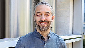 Sven Ramelow, a man with long gray hair and a beard, smiles at the camera. He is wearing a gray shirt and standing in front of a bright, modern wall. His face radiates friendliness and openness.