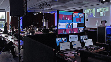 Hybride ISLC2021-Konferenz, im Vordergrund sind die Monitore zu sehen, über die Teilnehmende online zugeschaltet werden und im Hintergrund eine Vor-Ort-Präsentation mit Teilnemenden in Präsenz