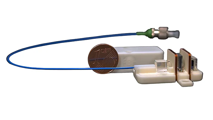 The image shows a ceramic magnetometer measuring head. The measuring head is shown open and consists of two copper coils and small optical elements mounted on a white ceramic substrate. An optical fiber with a blue housing is connected to it. A cent coin is placed next to the measuring head for size comparison.