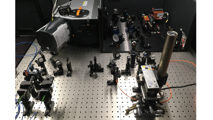 Wissenschaftliche Apparatur auf einer perforierten Metallplatte. Verschiedene optische Instrumente, Halterungen und Kabel sind verteilt.