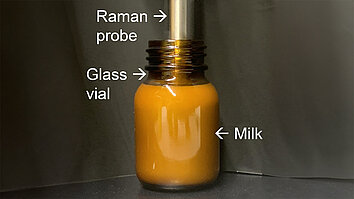 Metal Raman probe inside a brown glass vial. Labels are used to identify the key components: "Raman probe" points to the top part of the metal probe, "Glass vial" points to the neck of the transparent container, and "Milk" points to the liquid filling the vial.