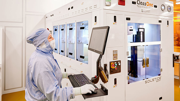 Eine Person in blauer Schutzkleidung bedient einen Computer in einem technischen Labor. Im Hintergrund sind Geräte mit transparenten Türen sichtbar.