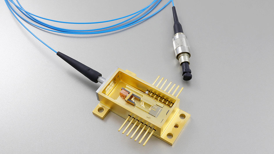 Gold-colored electronic component with multiple pins on a base. It contains an enclosed section with internal parts visible. Two blue optical fibers connect to the component, one ending in a round connector.