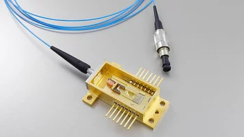 Image shows a gold-colored electronic component with multiple pins on a base. It contains an enclosed section with internal parts visible. Two blue optical fibers connect to the component, one ending in a round connector.