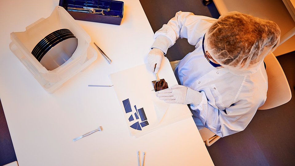 Eine Aufnahme von oben, die eine junge Frau zeigt, die gerade ein Waferstück in einzelne Chips zerteilt. Auf dem Tisch steht auch noch ein Behälter mit ganzen Wafern.