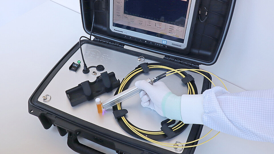 Tragbares Raman-Messsystem in einem geöffneten Schutzkoffer mit integriertem Display, Glasfaserkabeln und einer von einer behandschuhten Hand gehaltenen Optode über einem kleinen Flüssigkeitsröhrchen.