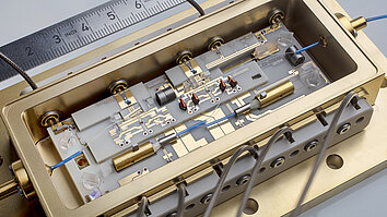Das Bild zeigt ein präzise konstruiertes photonisches Modul in einem goldfarbenen Gehäuse. Es enthält mehrere Komponenten, darunter Drähte, Linsen und Leiterplatten. Ein Lineal ist im Hintergrund sichtbar, um die Maßstäbe der technischen Details zu verdeutlichen.