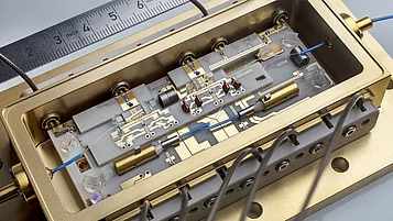 The image shows a precisely constructed photonic module in a gold-colored housing. It contains several components, including wires, lenses, and circuit boards. A ruler is visible in the background to illustrate the scale of the technical details.
