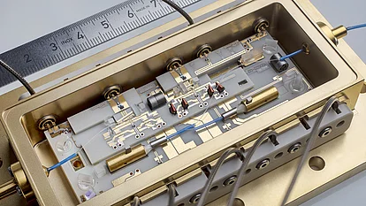 The image shows a precisely constructed photonic module in a gold-colored housing. It contains several components, including wires, lenses, and circuit boards. A ruler is visible in the background to illustrate the scale of the technical details.