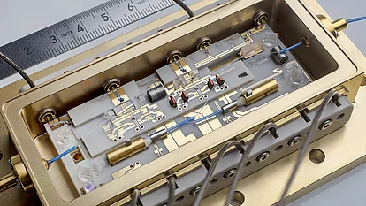 Das Bild zeigt ein präzise konstruiertes photonisches Modul in einem goldfarbenen Gehäuse. Es enthält mehrere Komponenten, darunter Drähte, Linsen und Leiterplatten. Ein Lineal ist im Hintergrund sichtbar, um die Maßstäbe der technischen Details zu verdeutlichen.