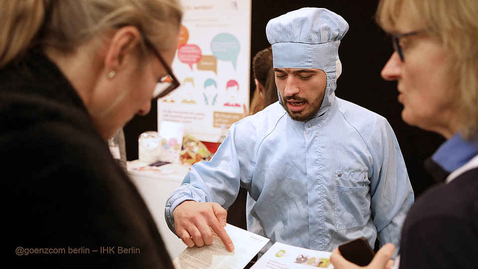 Ein Mitarbeiter vom FBH erläutert Interessenten eine Broschüre an einem Messestand.