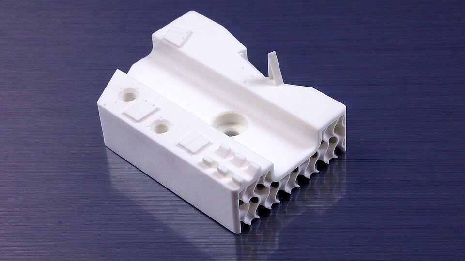 White ceramic substrate with several holes, raised features, and honeycomb-like openings at the front, resting on a dark reflective surface.