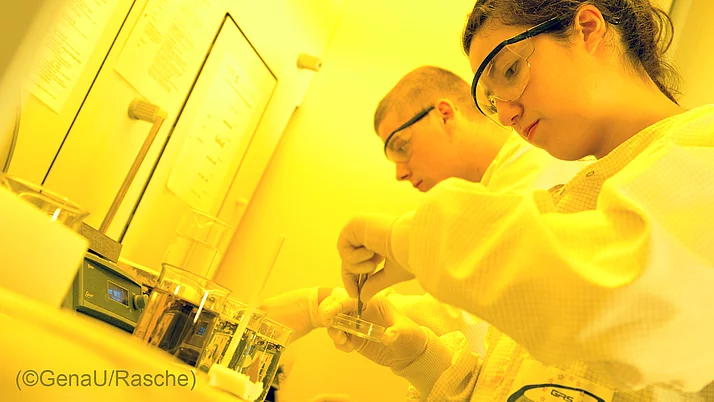 Zwei Personen in Laborkleidung arbeiten an einem Experiment. Die Frau hält eine Petrischale, während der Mann neben ihr schaut.