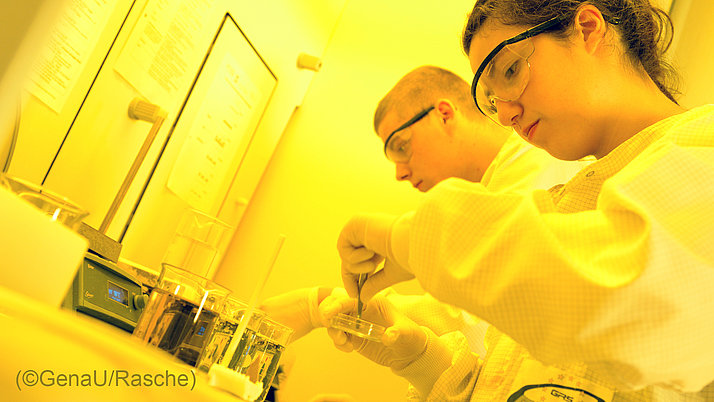 Zwei Personen in Laborkleidung arbeiten an einem Experiment. Die Frau hält eine Petrischale, während der Mann neben ihr schaut.