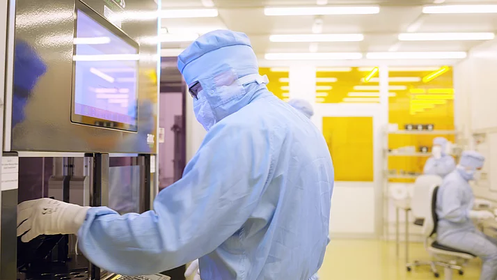 Videostill aus dem FBH-Reinraumfilm Mitarbeitende in vollständiger Reinraum-Schutzkleidung bedienen eine Anlage im hochmodernen Reinraumlabor des Ferdinand-Braun-Instituts. Im Hintergrund arbeiten weitere Personen an Tischen . Auf dem Bild steht der Text „2.000 m² cleanroom“.