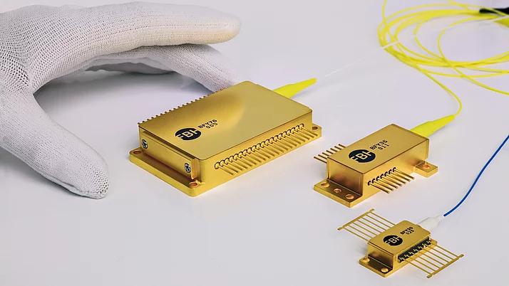 Eine Hand in einem weißen Handschuh hält mehrere goldene Diodenlasermodule, darunter ein größeres und zwei kleinere Module mit Anschlüssen und gelben Kabeln. 