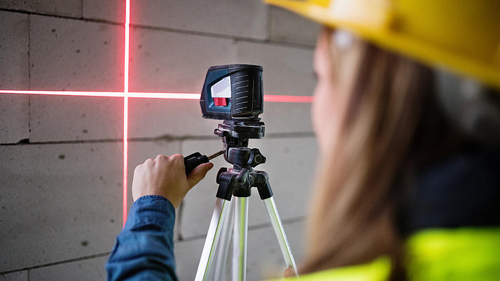 A person wearing a yellow safety helmet operates a laser device that is projected onto a wall. The device stands on a tripod, while a red laser beam creates a horizontal and vertical line on the wall.