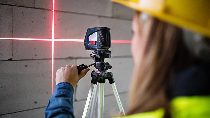A person wearing a yellow safety helmet operates a laser device that is projected onto a wall. The device stands on a tripod, while a red laser beam creates a horizontal and vertical line on the wall.
