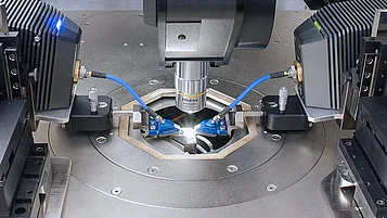 Microscopic measurement setup with a central objective above a sample. Two blue probes are positioned from the left and right toward the sample, with additional equipment surrounding the measurement area.