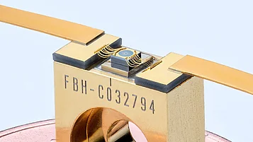 Close-up of a gold-colored component package marked “FBH-C032794.” A small blue circular element on top is connected to contact pads by several curved gold wires, with wide metal contacts extending to the left and right.
