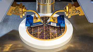 Das Bild zeigt eine hochpräzise Messapparatur, die zwei goldene Probenaufnahmegeräte über einer CPU-Wafer-Plattform platziert. Die Wafer-Oberfläche ist mit komplexen, strukturierten Mustern bedeckt, die in der Halbleiterfertigung verwendet werden.