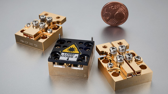 Three gold-colored components are arranged on a silver surface. The central component bears a black label with the words “Laser class 4”. There is a 1-cent coin in the background.