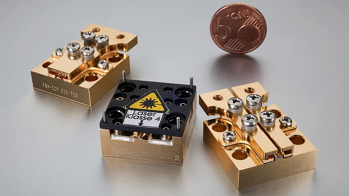 Three gold-colored components are arranged on a silver surface. The central component bears a black label with the words “Laser class 4”. There is a 1-cent coin in the background.