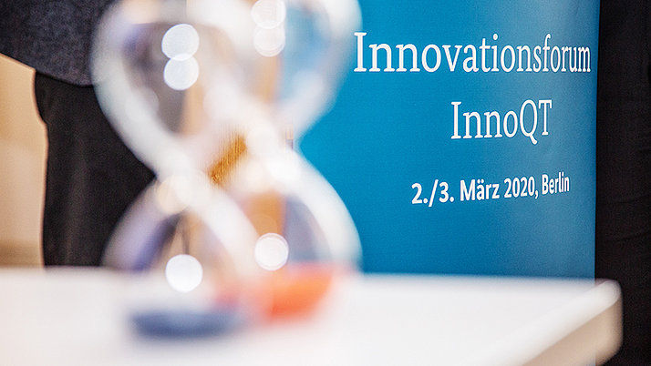 Zwei Sanduhren im Vordergrund. Im Hintergrund steht ein Banner mit der Aufschrift „Innovationsforum InnoQT, 2./3. März 2020, Berlin“.