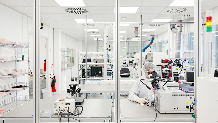 Ein Labor mit modernen Geräten. In der Mitte sitzt eine Person in Schutzkleidung mit einer Schutzbrille und arbeitet an einem Mikroskop.