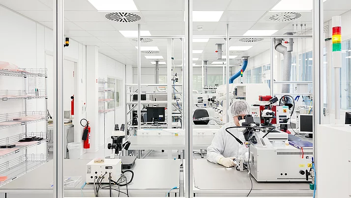 Ein Labor mit modernen Geräten. In der Mitte sitzt eine Person in Schutzkleidung mit einer Schutzbrille und arbeitet an einem Mikroskop.
