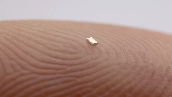 Ein winziges, rechteckiges Bauteil liegt auf der Hautoberfläche eines Fingers. Die Fingerabdrücke sind deutlich sichtbar und zeigen die Textur der Haut.