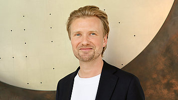 Portraitfoto von Markus Krutzik, eines lächelnden Mannes mit blonden, kurzen Haaren. Er trägt ein schwarzes Sakko über einem weißen T-Shirt und steht vor einer abstrakten, metallischen Wand. Die Hintergrundstruktur ist glänzend und hat eine harmonische, runde Form.