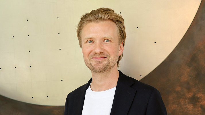 Portraitfoto von Markus Krutzik, eines lächelnden Mannes mit blonden, kurzen Haaren. Er trägt ein schwarzes Sakko über einem weißen T-Shirt und steht vor einer abstrakten, metallischen Wand. Die Hintergrundstruktur ist glänzend und hat eine harmonische, runde Form.