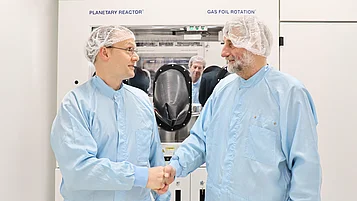 Zwei Personen in blauer Schutzkleidung schütteln sich die Hände vor einer technischen Anlage. Im Hintergrund sind mehrere Personen zu sehen.