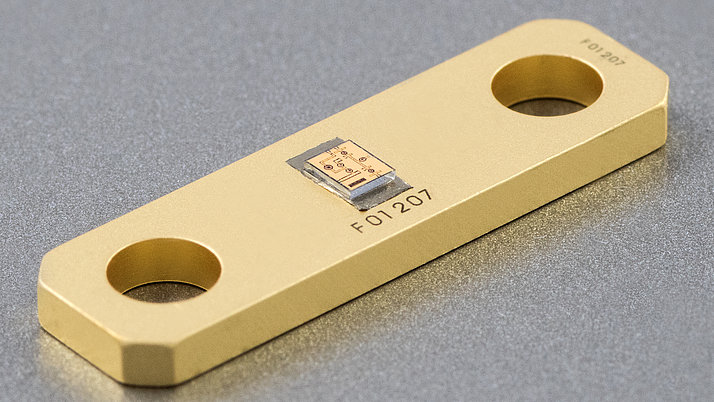 A gold-colored electronic component with two circular holes at each end. It features a rectangular chip affixed to its center, displaying printed circuit patterns.