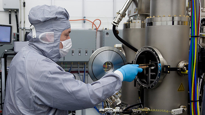 A person in a protective suit and gloves is working on a scientific device. They are holding a tool that is inserted into a metal container connected to various cables and hoses.