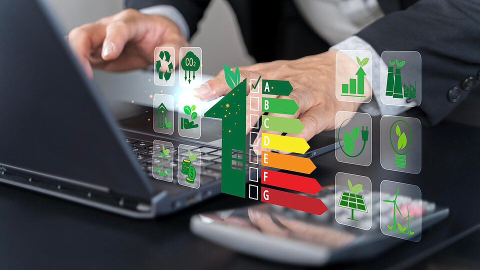 Eine Person arbeitet an einem Laptop, es sind nur die Hände und Arme zu sehen. Im Vordergrund ist ein Energieeffizienzdiagramm zu sehen, das von A bis G reicht. Neben dem Diagramm sind Symbole für Umweltaspekte wie Recycling, Solarenergie und Nachhaltigkeit dargestellt. Die Darstellung betont den Fokus auf energetische Effizienz und Umweltbewusstsein.