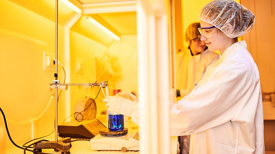 [Translate to English:] A young woman stands in a laboratory holding a wafer piece in a beaker of acid to etch away areas not covered with resist.