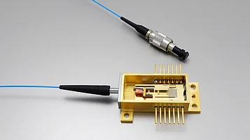 Goldene modulare Laserdiode auf einer Basisplatte, verbunden mit einem blauen Lichtwellenleiter. Der Wellenleiter und eine angeschlossene Stecker-Komponente sind ebenfalls sichtbar.