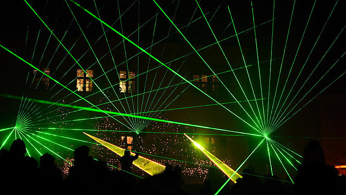 Green laser beams pierce the darkness, shining in different directions and creating a dynamic play of light. In the background, festive lights and shadows of people watching the spectacle can be seen.