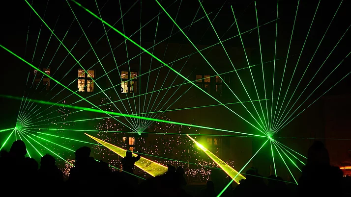 Green laser beams pierce the darkness, shining in different directions and creating a dynamic play of light. In the background, festive lights and shadows of people watching the spectacle can be seen.