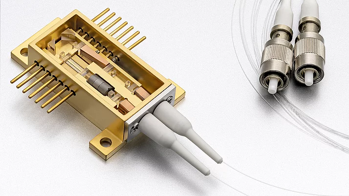 Das Bild zeigt ein geöffnetes Verstärker- und Frequenzkonvertermodul. Im Inneren befindet sich ein Mikroisolator, ein Halbleiterverstärker und ein nicht-linearer Kristall zur Frequenzverdopplung. Rechts sind zwei Glasfasern mit metallischen Steckern zu sehen. Der untere ist der Fasereingang, der obere der Faserausgang.