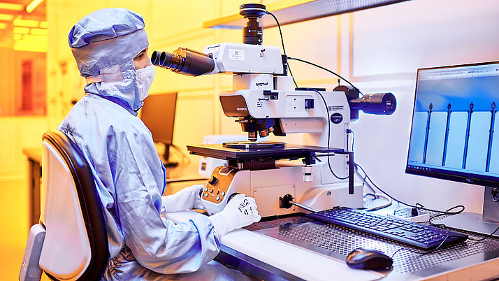 A laboratory technician, dressed in protective clothing, operates a microscope. The microscope is focused on a sample placed on its stage, while a computer monitor displays related data. 