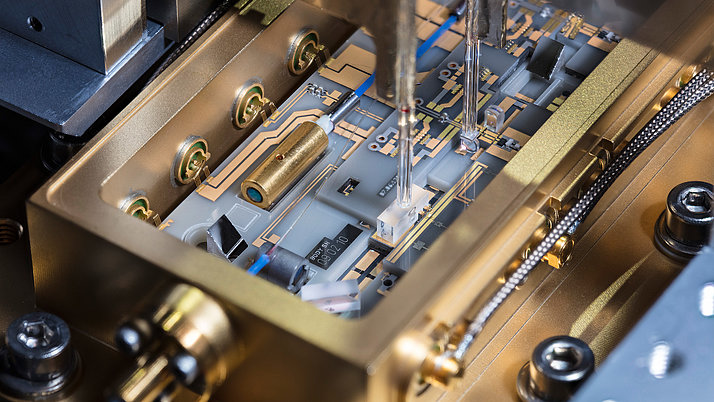 Nahaufnahme einer Leiterplatte in einem goldenen Gehäuse, auf der Kontaktstellen, goldene Drähte und elektronische Komponenten sichtbar sind. Eine feine Nadel ist in das Gehäuse eingeführt.
