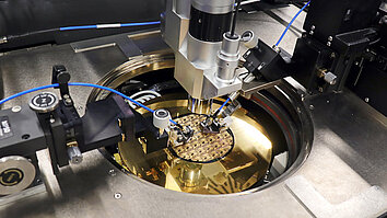 Das Bild zeigt eine präzise Maschine zur Waferbearbeitung in einem Labor. Im Mittelpunkt ist eine goldene Waferplatte zu sehen, während zwei Roboterarme aktiv mit der Wafer-Oberfläche interagieren. Die technische Ausrüstung deutet auf Fortschritte in der Halbleiterfertigung hin.