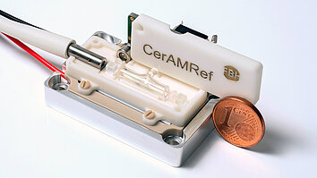 Micro-integrated frequency reference module on a silver base, consisting of white plastic parts, a metal cylinder, and connectors. A cent coin is placed next to it for size comparison. The system is labeled “CerAMRef”.