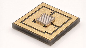 Das Bild zeigt einen mikroelektronischen Chip auf einem rechteckigen Trägermaterial. Der Chip hat eine goldene Oberfläche mit einem zentralen, kleineren Element, das in einem Quadrat angeordnet ist. Die Struktur besteht aus feinen Linien und Mustern, die für elektronische Anwendungen typisch sind.